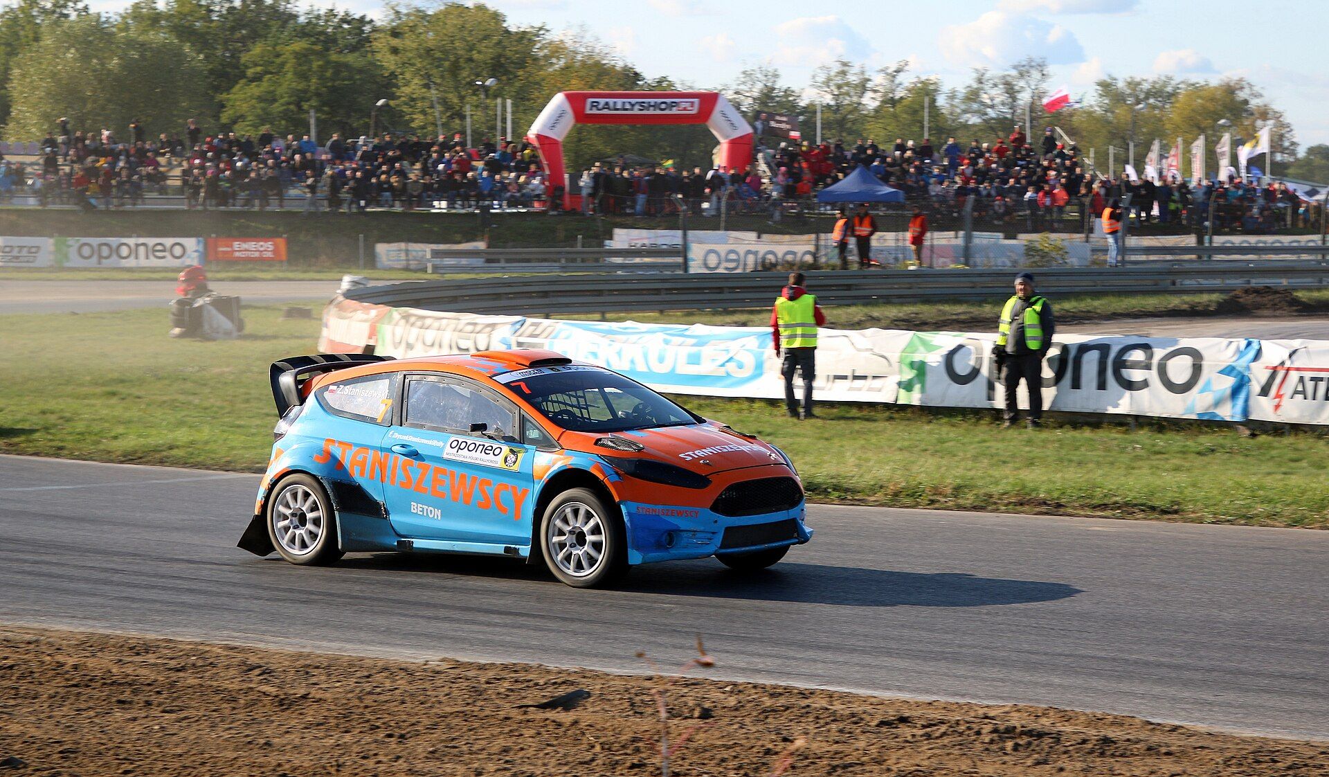 Samochód rallycrossowy na asfalcie toru MotoPark Toruń, za bandą trybuna wypełniona kibicami